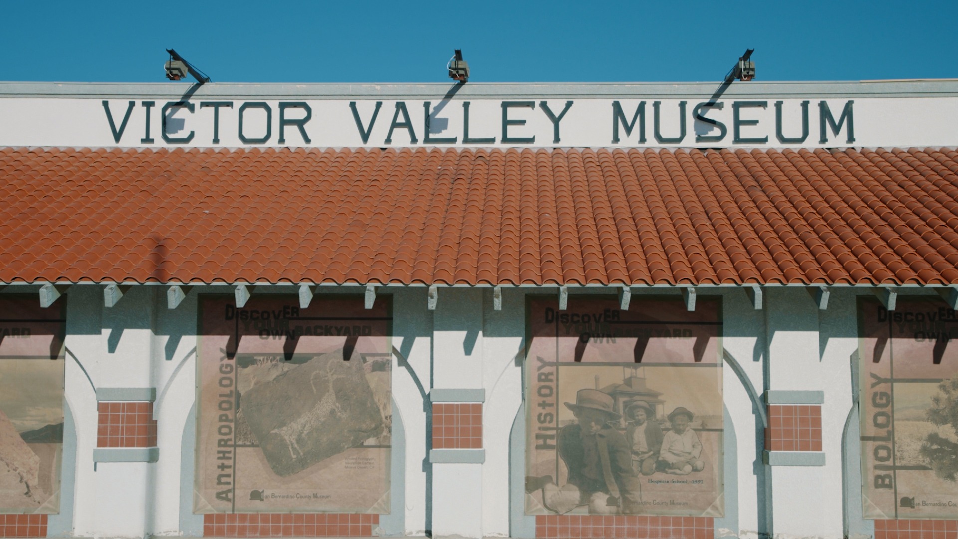 The image shows the Victor Valley Museum's exterior with a red-tiled roof and a large sign. Below are display panels with images related to Anthropology, History, and Biology.